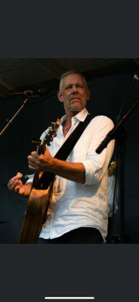 Person i vit skjorta spelar akustisk gitarr på scen med mikrofoner runt sig.