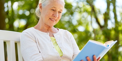 Äldre kvinna med vitt hår sitter utomhus på en bänk och läser en bok, omgiven av grönska.