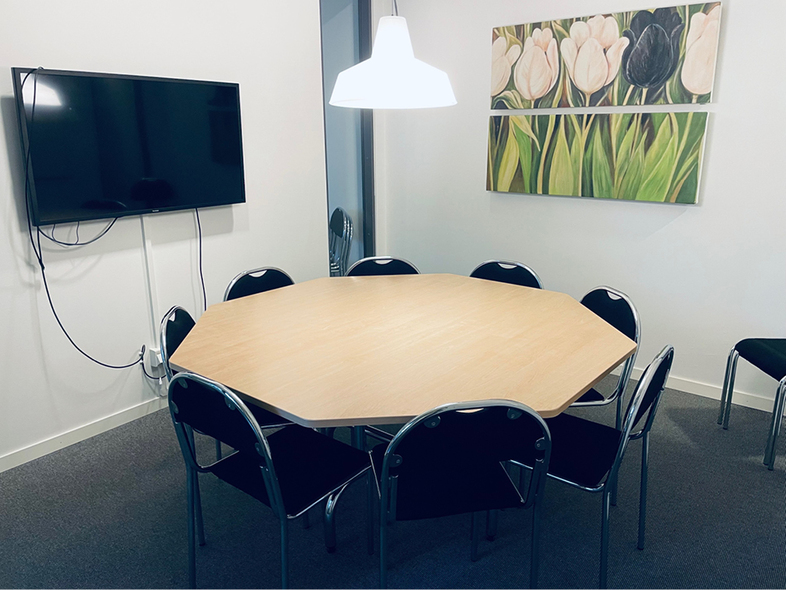 Ett mötesrum med ett åttakantigt bord, åtta stolar, en TV på väggen och en tavla med blommor.