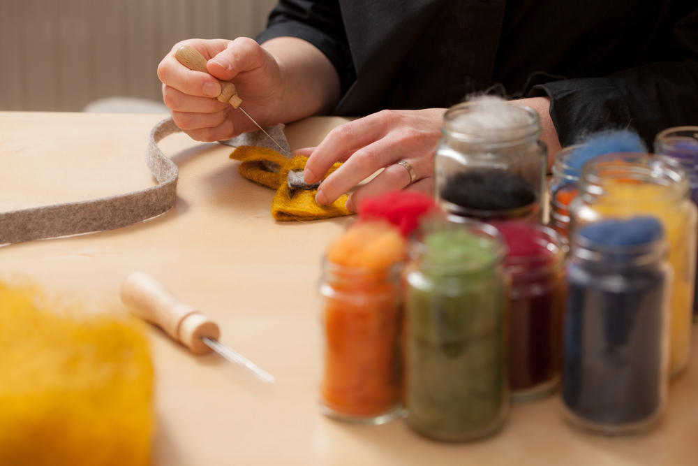 En person arbetar med nåltovning vid ett bord, omgiven av burkar med färgad ull.