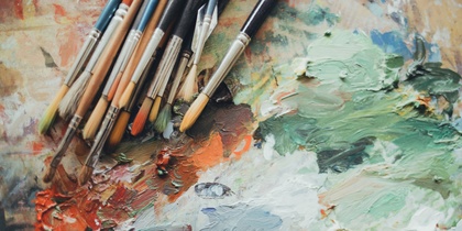 Flera penslar ligger på en färgpalett med blandade oljefärger i grönt, vitt och orange.