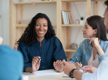 Fyra personer sitter runt ett bord och har ett möte, en kvinna i fokus ler och pratar.