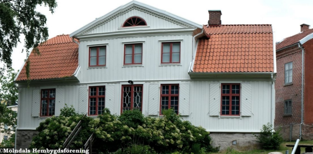 Gammalt vitt trähus med rött tegeltak och röda fönster, omgiven av buskar och träd.