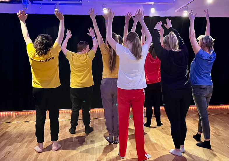 Sju personer står med ryggen mot kameran och armarna uppsträckta i ett upplyst rum med trägolv.