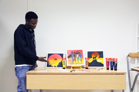 En man står vid ett bord med tre färgglada målningar och tuber med akrylfärg.