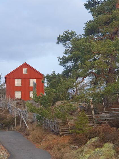 Rött trähus vid en grusväg, omgiven av träd och ett trästaket under en mulen himmel.