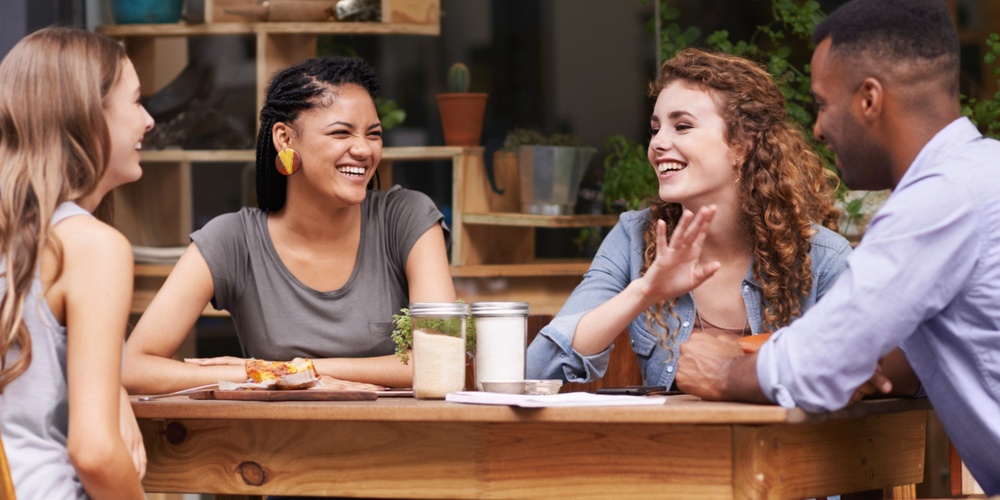 Fyra personer sitter vid ett träbord utomhus och samtalar, med växter i bakgrunden.