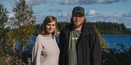 En kvinna och en man står utomhus vid en sjö med skog i bakgrunden under en solig dag.