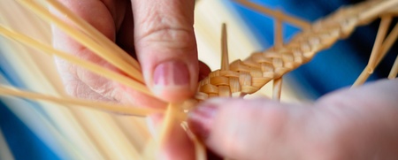 Närbild av händer som flätar tunna strån i ett hantverksarbete.