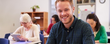 Leende man sitter vid skolbänk i klassrum, skriver i anteckningsbok. Andra vuxna studenter i bakgrunden.
