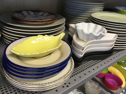 Olika staplade tallrikar och fat i olika former och färger på en hylla i ett kök eller butik.