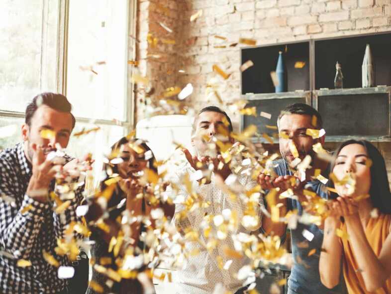 En grupp personer blåser guldkonfetti från händerna mot kameran.