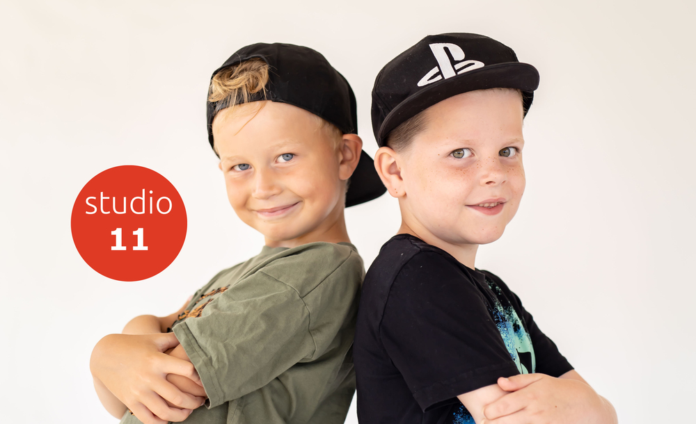 Två leende pojkar står rygg mot rygg med armarna i kors, båda bär svarta kepsar. Texten 'studio 11' syns till vänster.