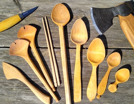 Olika handgjorda träredskap, som skedar och spatlar, ligger på ett träbord tillsammans med en kniv och en yxa.
