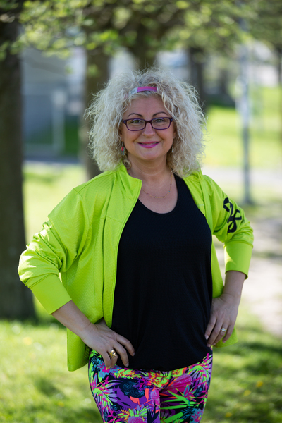 Person med lockigt blont hår, gul jacka och färgglada byxor står utomhus i en park.