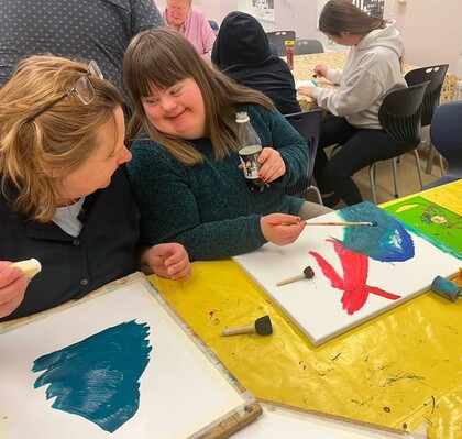 Två personer målar tillsammans vid ett bord, ler mot varandra och håller i penslar och färg.