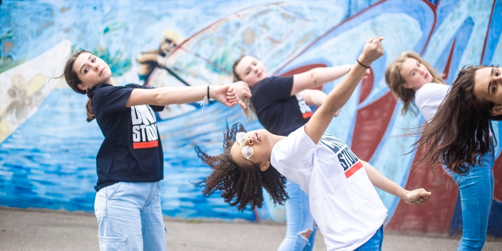 Fyra unga personer dansar energiskt framför en blå graffitivägg utomhus.