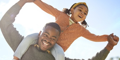 En glad man bär en leende flicka på sina axlar utomhus under en solig dag.