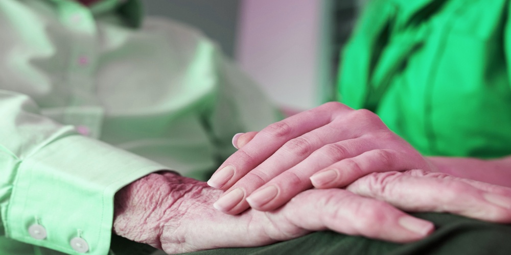 En yngre person håller en äldre persons hand i en stödjande gest. Båda bär gröna kläder.