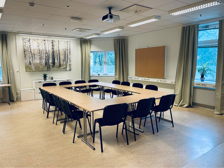 Ett tomt konferensrum med U-formade bord, svarta stolar och stora fönster med gröna gardiner.