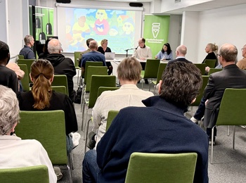 En grupp människor sitter på gröna stolar i ett konferensrum och lyssnar på en paneldiskussion med presentation på skärm.