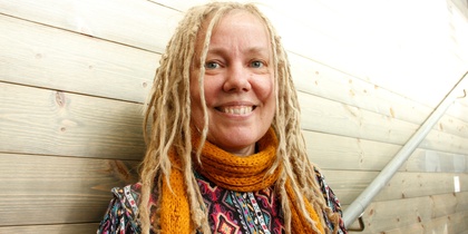 Leende kvinna med blont dreadlockshår och orange halsduk mot en trävägg.