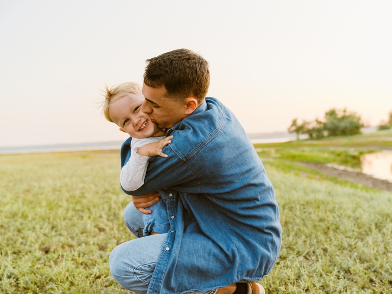 En vuxen och ett barn kramas och skrattar utomhus på en grön äng vid solnedgången.