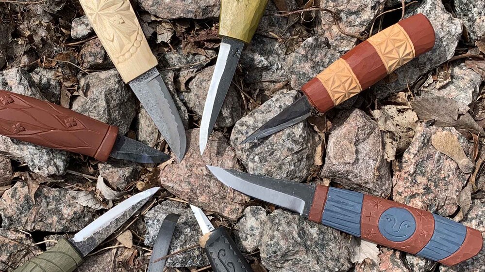 Sju traditionella knivar med snidade träskaft ligger i en cirkel på stenar utomhus.