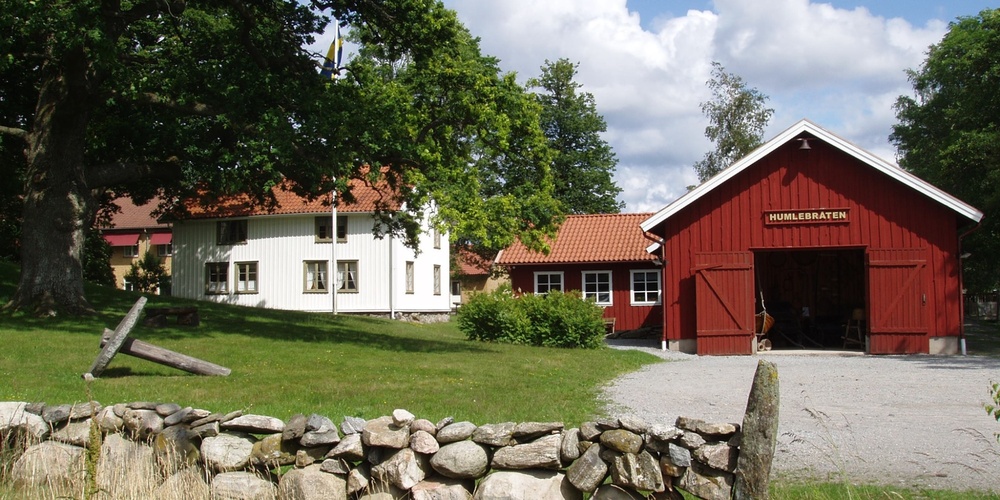 Gårdsmiljö med röd lada, vit byggnad, stengärdsgård och grönskande träd under blå himmel.