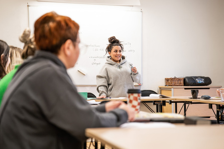 En kvinna står framför en whiteboard och pratar med en grupp personer i ett klassrum.