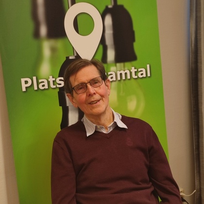 Man med glasögon sitter vid ett bord framför en grön skylt med texten 'Plats för samtal'.