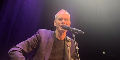 Man i kostym spelar akustisk gitarr och sjunger i mikrofon på scen under stark belysning.