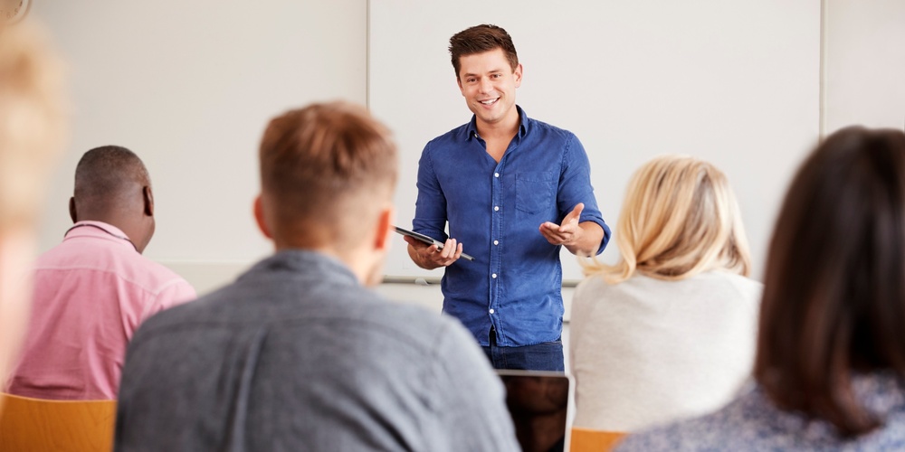 En man står framför en grupp och håller en presentation i ett klassrum.