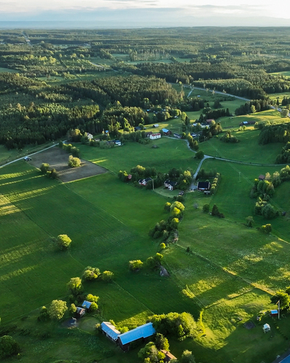 Flygfoto över ett grönt landskap med åkrar, skog och utspridda hus i kvällsljus.