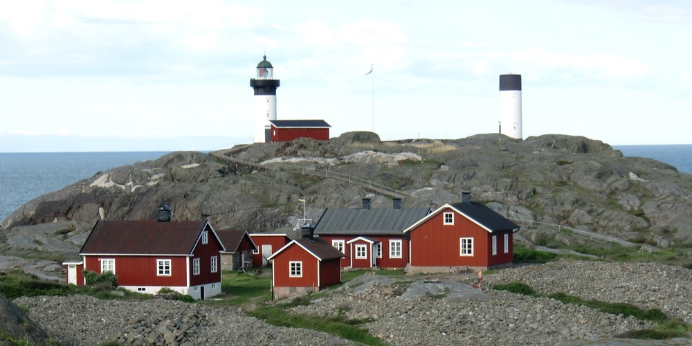 Flera röda stugor och en fyr på en klippig ö vid havet, med en segelbåt i bakgrunden.