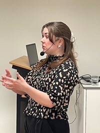 Kvinna med headset håller presentation vid talarstol, bär mönstrad blus och har långt hår.