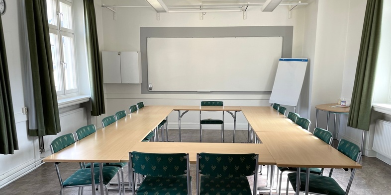 Ett tomt mötesrum med U-formade bord, gröna stolar, whiteboard och blädderblock.