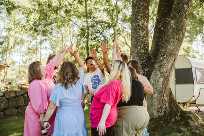 En grupp människor står i en ring utomhus och sträcker upp händerna tillsammans under träd.