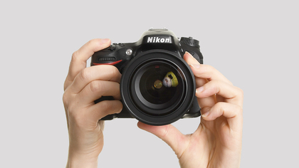 Två händer håller en Nikon-systemkamera mot en ljus bakgrund.
