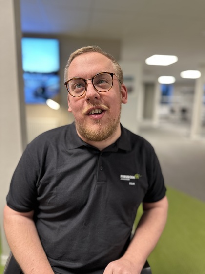 Man med glasögon och skägg sitter inomhus i en modern kontorsmiljö, bär svart pikétröja.