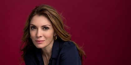 Person med långt brunt hår och mörkblå skjorta mot vinröd bakgrund.