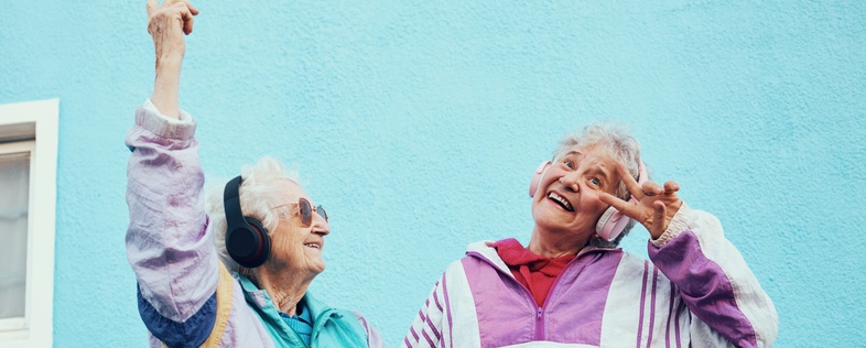 Två glada äldre kvinnor i färgglada träningsjackor poserar och dansar framför en blå vägg.