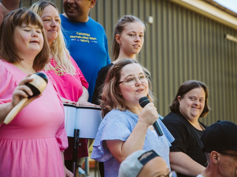 En grupp människor, flera med mikrofon och musikinstrument, står utomhus och sjunger tillsammans.