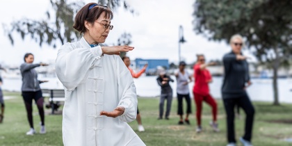 En grupp människor utövar tai chi utomhus i en park, med en kvinna i vit dräkt i förgrunden.