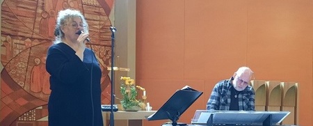 En kvinna sjunger i mikrofon och en man spelar keyboard på en liten scen i en kyrka med orange väggar.