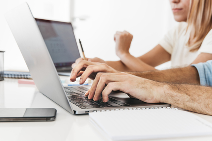 Två personer arbetar vid en bärbar dator med anteckningsblock och mobiltelefon på ett vitt skrivbord.