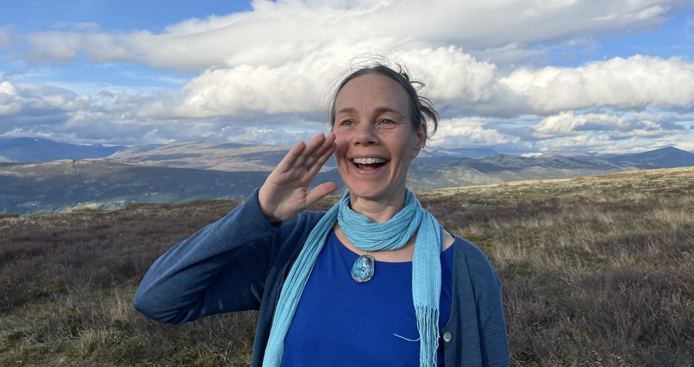 Person står utomhus på fjäll med blå himmel och moln, klädd i blått och turkos halsduk.
