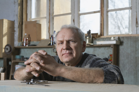 Äldre man med grått hår sitter vid ett arbetsbord i ett ljust, rustikt rum och tittar mot kameran.