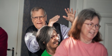 Fyra äldre personer skrattar och rör sig tillsammans inomhus, i en ljus och glad miljö.