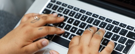 Två händer med ringar och tatueringar skriver på ett svartvitt tangentbord på en bärbar dator.